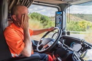A man in an orange shirt drives a truck while rubbing his eye, appearing tired, with a highway and greenery visible outside the window.