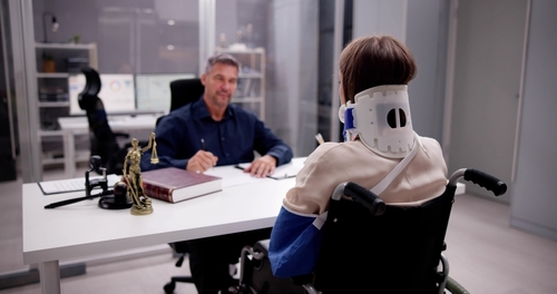 A person in a wheelchair with a neck brace and arm sling sits across from a professional at a desk in an office setting.