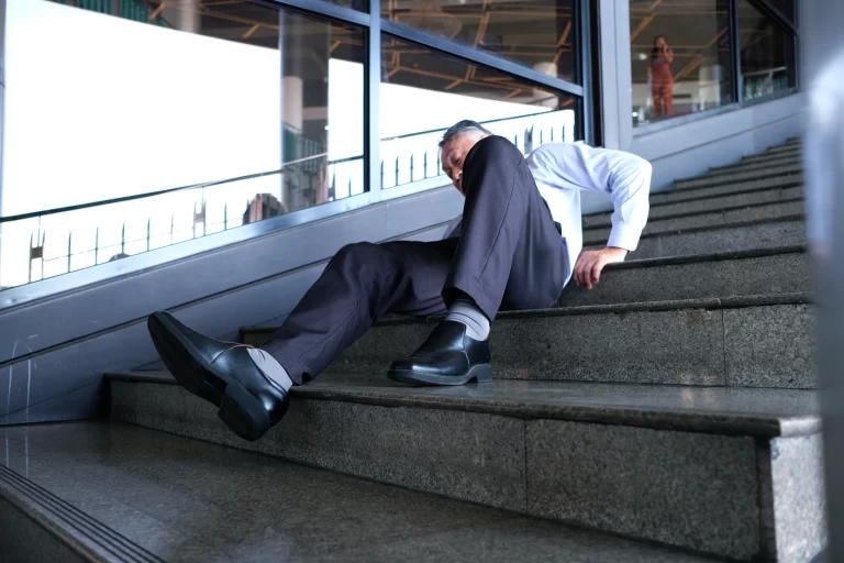 A man in formal attire is lying on a set of stairs, appearing to have fallen, with one leg bent and his hand on the step for support.