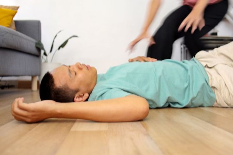 A person lies unconscious on a wooden floor while another person kneels nearby, possibly to assist.