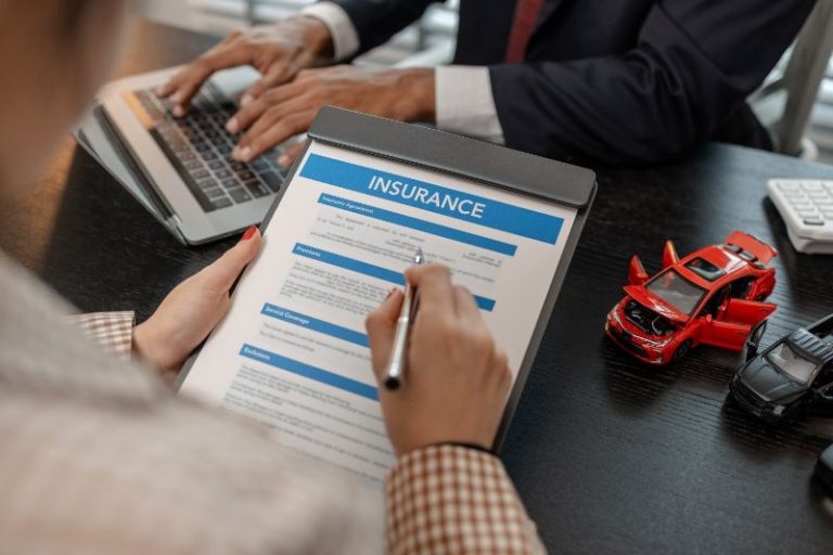 Person holding an insurance form on a clipboard while another person types on a laptop; toy cars are on the desk, suggesting a car insurance context.