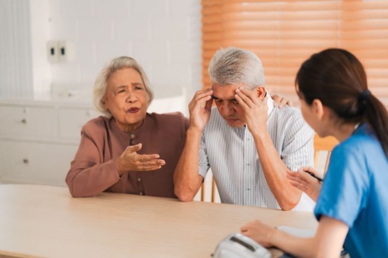 An elderly man sits at a table with his head in his hands, an elderly woman gestures beside him, and a healthcare professional listens across from them.