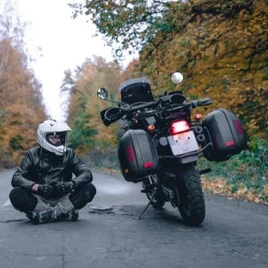 A person in motorcycle gear sits on a road next to a parked touring motorcycle; autumn trees line both sides of the road.