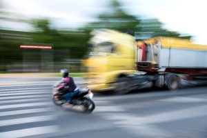 A motorcycle and a yellow truck pass each other at a crosswalk, both in motion with a blurred background indicating speed.
