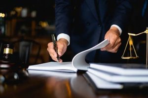 A person in a suit writes in a document with a pen, surrounded by books, a gavel, and a balance scale on a desk.