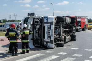 A large truck lies on its side at an intersection while firefighters and emergency vehicles are present at the scene.