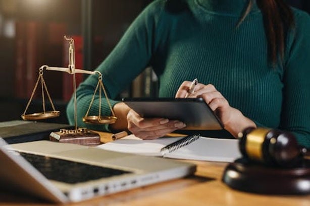 A person in a green sweater works at a desk with a laptop, tablet, scales of justice, gavel, and notebook.