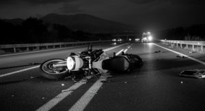 A motorcycle lies damaged on a dark, empty road with debris scattered around; emergency vehicle lights are visible in the background.