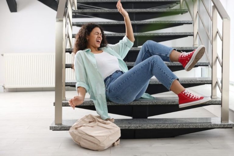 A woman in jeans and red sneakers appears to be falling backward on indoor stairs, with a beige bag on the floor nearby.
