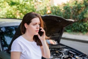 A woman stands in front of a car with the hood open, looking concerned while talking on her phone.