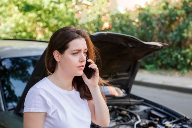 A woman stands in front of a car with the hood open, looking concerned while talking on her phone.
