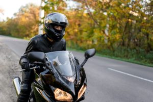 Person in black motorcycle gear and helmet riding a black sport bike on a road with trees in the background.