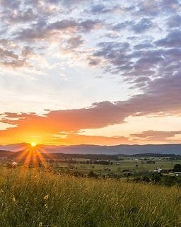 Sunrise over a grassy field with scattered houses, distant hills, and a sky filled with colorful clouds.