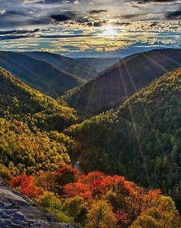 Sunset over a valley with forested hills; trees display green, yellow, and orange foliage, and rays of sunlight stream through clouds above.