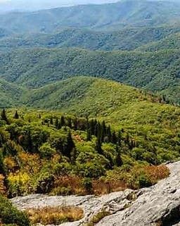 A panoramic view of rolling green hills and forested mountains with a rocky foreground under a cloudy sky.