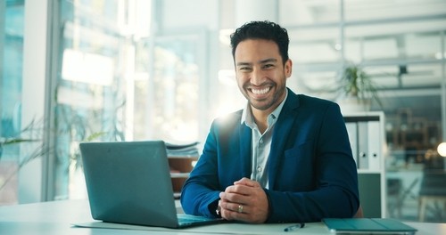 Smiling man in a blue suit sits at a desk with a laptop in a bright office environment.