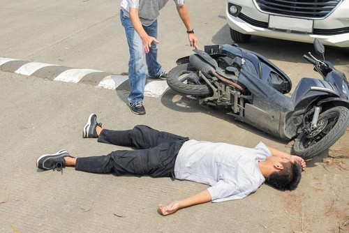 A person lies on the ground next to a fallen motorcycle in front of a car, while another person stands nearby with hands outstretched.