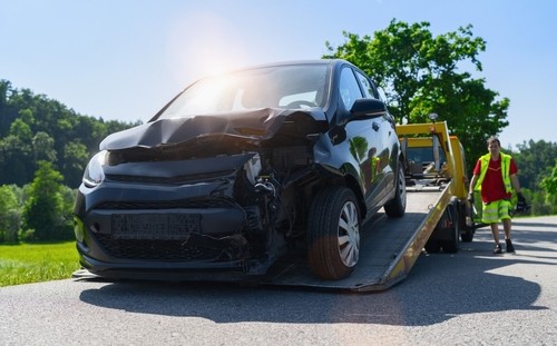 A black car with a damaged front end is being loaded onto a tow truck on a sunny day, with a worker standing nearby.