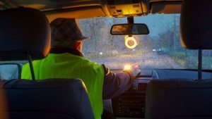 Person in a high-visibility vest sits in a car during rain, with hand on the dashboard; view is from the back seat looking out a wet windshield at dusk.