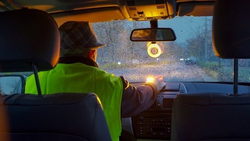 Person in a high-visibility vest sits in a car during rain, with hand on the dashboard; view is from the back seat looking out a wet windshield at dusk.