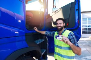 A man in a reflective vest stands by a blue truck and holds up a commercial driver’s license card, smiling at the camera.
