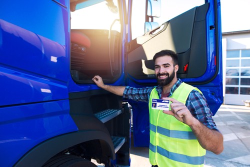 A man in a reflective vest stands by a blue truck and holds up a commercial driver’s license card, smiling at the camera.