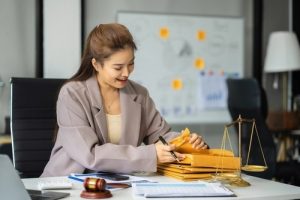A woman at a desk organizes folders, with a gavel, scales of justice, and documents in front of her in an office setting.