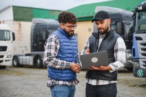 Two men standing outdoors by trucks, looking at a laptop. Both are wearing plaid shirts and vests.