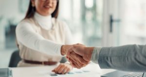 Two people shake hands across a desk in an office setting, with documents and a laptop visible on the table.