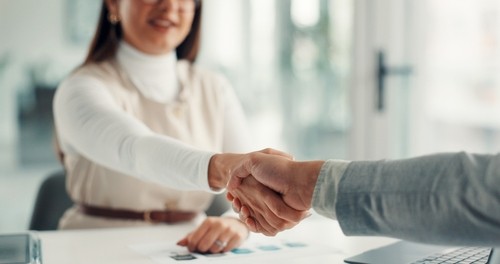 Two people shake hands across a desk in an office setting, with documents and a laptop visible on the table.