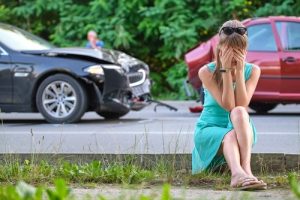A woman in a turquoise dress sits on a curb covering her face, while two damaged cars are seen on the road behind her after an apparent collision.