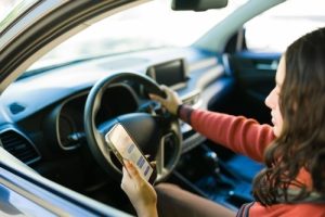 A woman sits in the driver’s seat of a car, holding a smartphone and looking at the screen while the vehicle appears to be stationary.