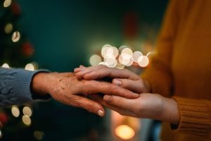 A younger person gently holding the hand of an older person, with blurred holiday lights in the background.