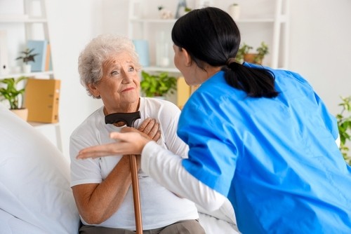 An elderly woman holding a cane sits on a bed while a caregiver in blue scrubs gestures and speaks to her in a bright, homey room.