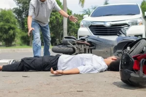 A person lies on the ground near a fallen motorcycle in front of a car, while another person stands nearby with arms outstretched.