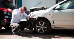 A man kneels next to a damaged white car after a rear-end collision with a black car, appearing to examine the damage.