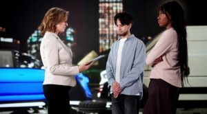 Three people stand outdoors near parked vehicles at night; one woman holds a clipboard and speaks to a man and another woman, who appear to be listening.