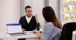 A man with a bandaged forearm discusses paperwork with a woman in an office; a laptop screen displays a health insurance claim form.