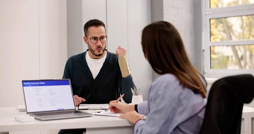 A man with a bandaged forearm discusses paperwork with a woman in an office; a laptop screen displays a health insurance claim form.