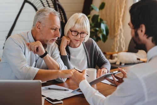 An older couple sits at a table with paperwork as a man shows them information on a tablet.