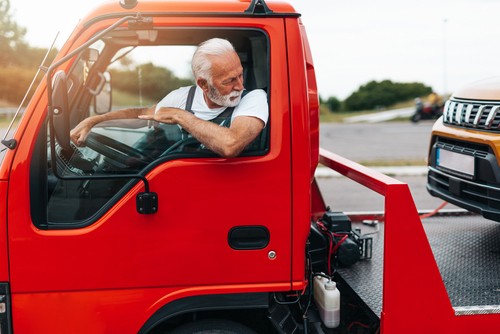 An older man with a white beard drives a red tow truck, looking backward as he tows an orange SUV.