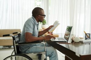 A man in a wheelchair works at a desk, holding documents and using a laptop in a bright, modern room.