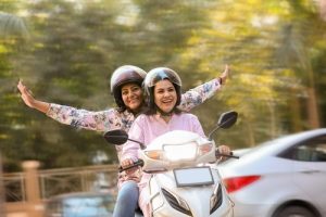 Two women wearing helmets ride a white scooter; the passenger extends her arms outward while both appear to be smiling, with blurred trees and a car in the background.